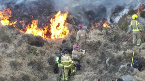 NIFRS Fire fighters at the scene of the fire in Bloody Bridge, County Down. Six of them can be seen in tan-coloured uniforms with bright yellow trim. They are standing in gorse which is not burning but in front of them there is gorse which is burning, orange flames are jumping up from it.