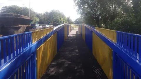 Shropshire Council A blue and yellow painted metal footbridge that runs parallel to a bridge with cars going over it. In the distance are traffic lights and a large roundabout 