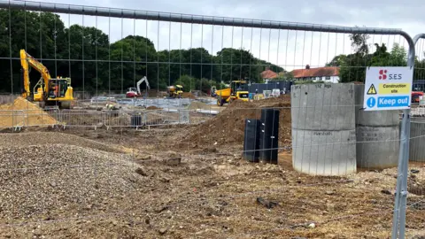 David Freezer/BBC There is a construction fence and behind it work is taking place on the building site. Not much has been done at this point, but in the background there are small construction vehicles.