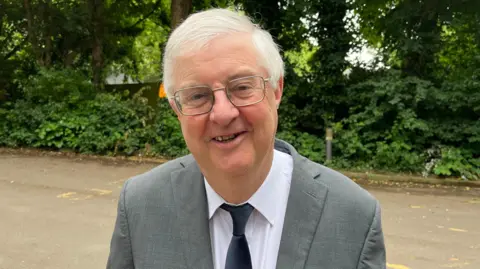A head and shoulders shot of Mark Drakeford. He has a grey suit, white shirt and blue tie. He has silver thin-rimmed glasses with short grey hair and smiles at the camera. There are bushes behind him. 