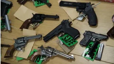 Northamptonshire Police Various pistols on a table. Some are black and one is grey. Some have green labels attached which say "SAFE". Some have light brown slips of paper attached.