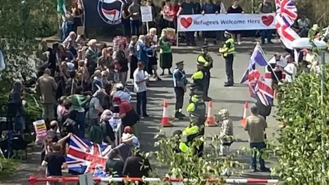 Protesters outside a hotel used to house asylum seekers on the edge of Exeter. Some are waving union flags and police are present in hi-vis uniforms.