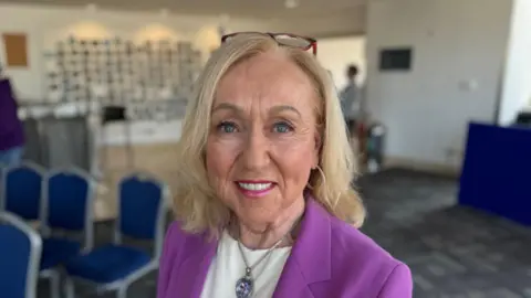 A woman wearing a purple blazer, white shirt, necklace and earrings is smiling at the camera. She has blonde hair and reading glasses resting on her head. Behind her are some blue chairs. 