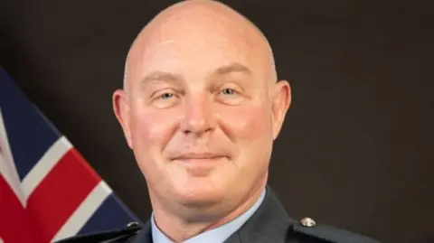 Royal Air Force A head and shoulders shot of Harv Smyth. He is bald man and is wearing the blue tunic and shirt of the RAF officer's uniform. To the lower left of the image behind him a union flag can be seen.