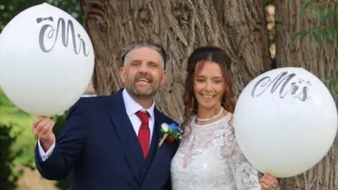 Celeste A groom and bride in their wedding outfits pictured in front of a tree, holding white balloons saying Mr and Mrs