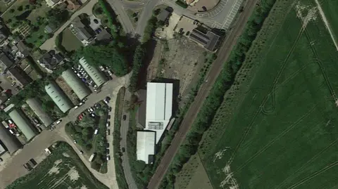Drone image of the sit with a white warehouse with fields on side and homes on the other side.