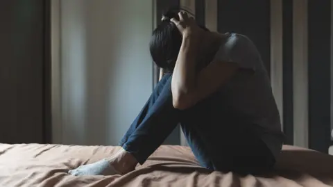 Getty Images A woman with dark hair, a grey T-shirt and jeans sits on a bed with her head in her hands
