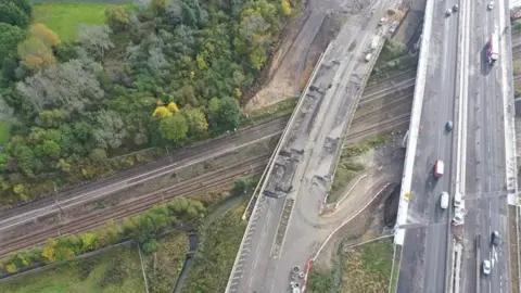 National Highways  The picture shows an aerial image of an old bridge, no longer in use with a new one next to it. Both cross a railway line  