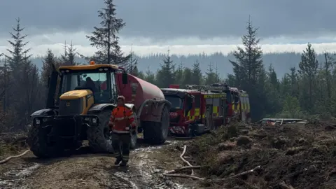 BBC Man walking in front of a tractor and fire engines in moorland.
