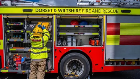 DWFRS A firefighter loading equipment into the side of a red and yellow fire engine. He is wearing a high-vis coat which reads "fire" on the back.