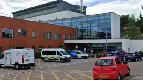 The outside of Pontefract Hospital which is a modern-looking building with brown tiles on one block and glass on another. There are three ambulances parked outside and a few cars.