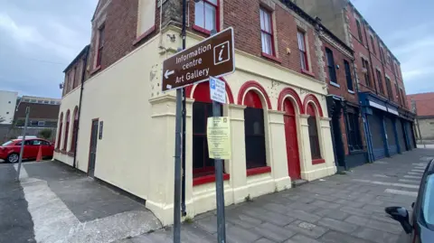 The exterior of a terraced building with the ground floor painted yellow with red paint around the window arches and doorway. A sign stands in front of the building pointing left reading "Information centre and Art Gallery".