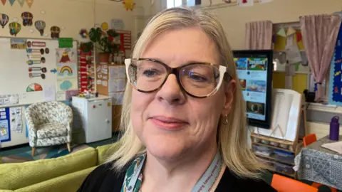 BBC Hilary Cunningham is in a classroom, she is wearing white and black glasses and a brown top. She has blonde hair. Behind her is a white board with decorations, a chair, a white fridge and an interactive board. The cream walls are covered with displays. 