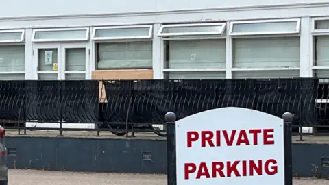 a private parking sign sits in front of a fence which has a set of windows behind it, one of which has been removed after being broken