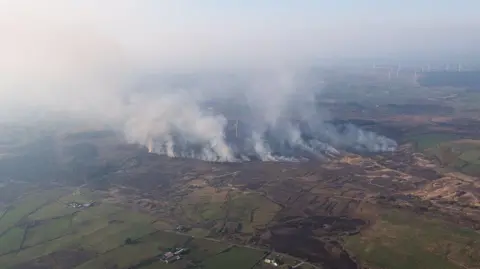 PSNI AIR SUPPORT An aerial image of a gorse fire in Ballygawley, smoke is billowing from the round with red lines of fire across fields. You can see white wind turbines around the smoke. 