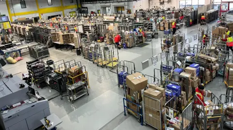 A large warehouse with high ceilings and bright lighting, filled with numerous shelves, carts, and packages. Several workers in high-visibility vests are seen throughout the space, organizing and moving items. The warehouse is well-organized with clear pathways and sections for different types of goods, depicting a busy and efficient logistics or distribution center.