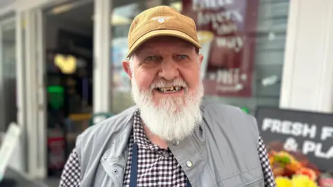 Robert Fox smiles, he has a long grey beard. He wears a yellow hat, checked brown shirt and grey gilet.