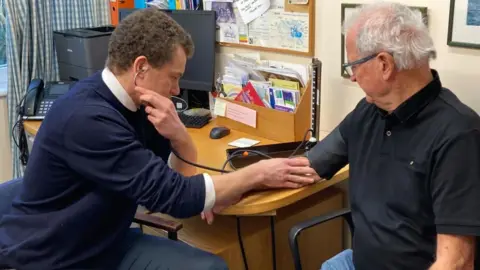 Two men are pictured seated. The man on the left is a doctor. He is taking the blood pressure of the man to the right. They in a doctor's surgery.