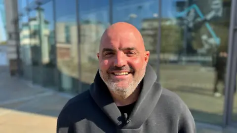 A man wearing a black hooded sweater is smiling into the camera. He has grey stubble and a bald head. Behind him is a large window. 