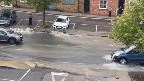 Water flows down a busy single lane road. Cars attempt to drive through it.