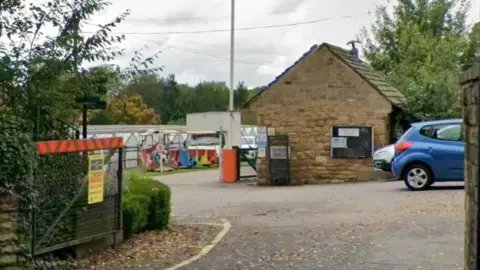 The picture shows the entrance to the site. The metal gate is open and several rows of greenhouses can be seen in the background.