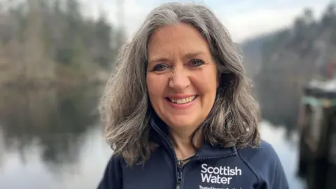 A woman with dark hair looks to the camera in front of Loch Katrine wearing a blue Scottish Water top 
