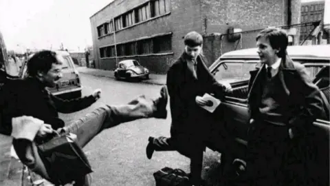 Grainy black-and-white photo of a young Bernard Sumner kicking his leg towards a young Ian Curtis. Rob Gretton is nearby, smiling and leaning against a car. They are standing in a street in front of a brick warehouse.