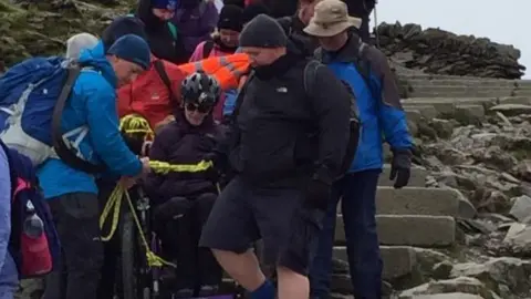Support Dogs Woman in wheelchair climbing a mountain with help of others