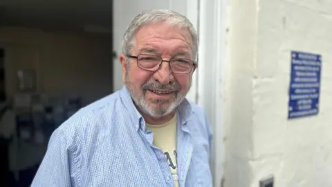 Alby Oldham stands by a white wall, he has short grey hair. He wears spectacles and a blue checked shirt, with a yellow T-shirt underneath.