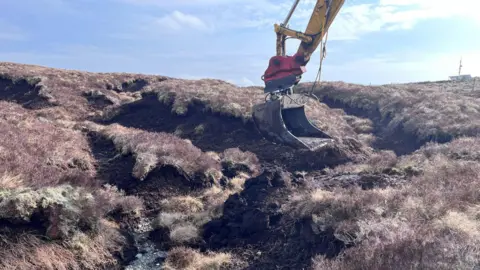 A digger's bucket hovers above exposed areas of peat which is being reshaped to protect it from the weather
