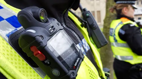 The image shows a close-up of a small black box attached to the hi-vis vest of a police officer. A red button on the side has the letters "REC" in white underneath.