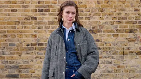 Female Topshop model wearing a dark grey winter coat over a dark blue pinstriped shirt and jeans, standing in front of a brick wall