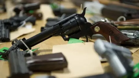 Northamptonshire Police Black pistols lying on a table. One is angled, resting on the end of the barrel and the bottom of the handle with the barrel in the air. There are green cards attached to each gun.