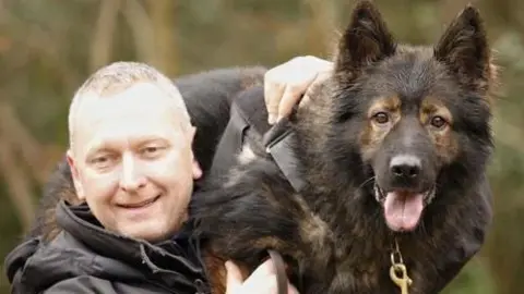 A blond man in a black jacket holding a Belgian Malinois on his left shoulder. The dog has a black lead with gold chain and its tongue is hanging out. The man is smiling. They are standing outside, with greenery out of focus behind.