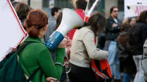 Getty Images A woman wearing a green jumper facing away from the camera and holding a megaphone in crowd of people at protest, some holding placards.