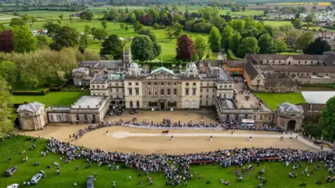 Stately home Badminton House with crowds in front of it and trees and fields behind it. It is a drone view up and looking down at the front of the house. 