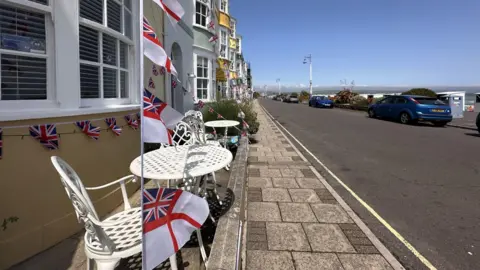 View from the pavement along a row of colourful Georgian townhouses on Weymouth seafront. The nearest building is painted yellow and has white tables and chairs outside and is decorated with bunting of the Royal Navy White Ensign flag. Cars are parked on the other side of the road and behind the cars is the beach and the sea.