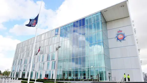 PA Media A blue flag with the Greater Manchester Police logo is flying at half mast outside a glass-fronted building which is the headquarters for the force. Two police officers are standing in front of the entrance. 