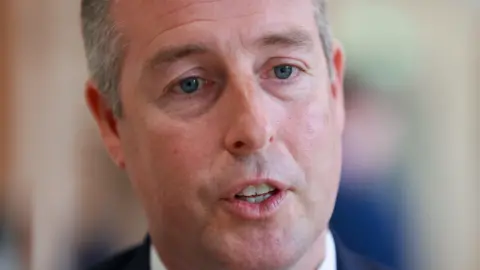 PA Media Paul Givens face can be seen, he is wearing a white shirt and navy blazer. The background is blurry. He has grey and black hair. 