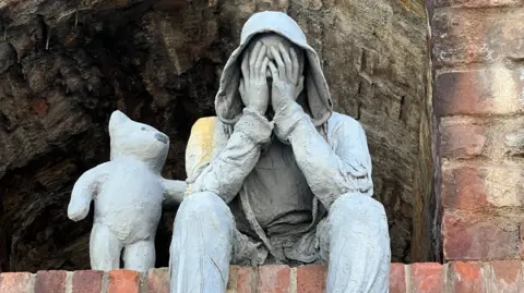 A close up view of a grey sculpture depicting a seated, hooded male figure with hands covering its face, accompanied by a bear-like companion offering comfort with a paw on the figure’s shoulder. 