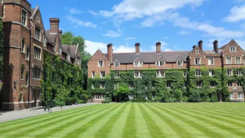 James Emmans/Geograph An external view of Selwyn College. It shows four storey red brick buildings at a right angle to each other, and their lower halves covered in green ivy. In front of them is a green lawn, mowed into stripes. Above is blue sky with some white clouds. 