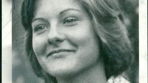 A black and white photo of a smiling young woman looking away from the camera. She is wearing a 1970s-style shirt with a geometric pattern and big wing collars.