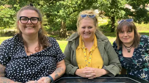 Three women sitting side-by-side on a park picnic bench. They are smiling at the camera and there are trees in dappled sunshine in the background.