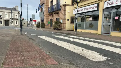 A section of road in Ramsey in front of the town hall, which sits next to a chip shop. You can see tired-looking pavements on both sides of the grey tarmac.
