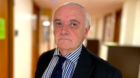 Chris Bown is looking directly at the camera and has a serious face. He has grey short hair, is wearing a navy suit jacket, navy and white striped shirt and navy and red tie. He is standing inside the hospital.