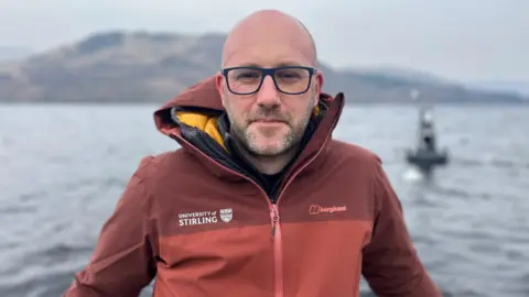 Professor Peter Hunter, in a two-tone red jacket and black-rimmed glasses looks at the camera in front of a buoy on Loch Katrine