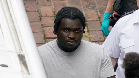 A man in handcuffs. He is being held by two men, one wearing a white short sleeved shirt and another wearing a black coat.