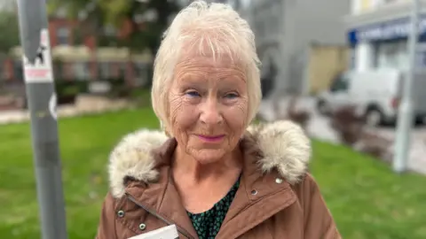 Winifred Whitehouse smiles, she has short blonde hair. She wears a brown coat with a furry collar and pink lipstick.
