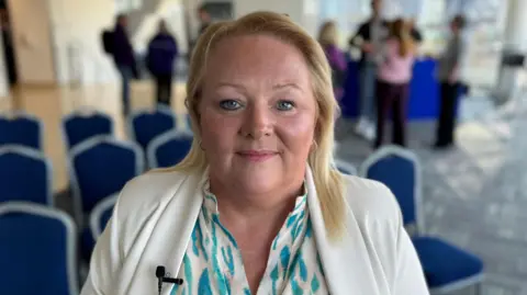 BBC A blonde haired woman, wearing a white blazer and blue patterned shirt is looking directly at the camera. Behind her are some blue chairs and people gathered in conversation. 