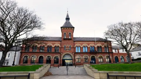 Hartlepool Borough Hall, an Italianate style building in red brick with stone dressings over each of two rows of 11 high-arched windows. In the centre there is a three-stage tower with a wide arched opening on the ground floor and steps leading up to the entrance.